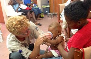 A volunteer kneeling by a woman with a baby, giving the baby some water.